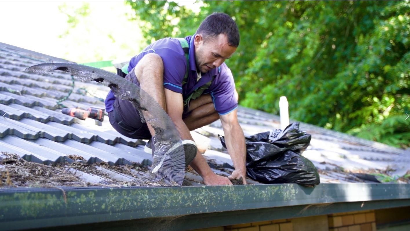 Worker kneeling on a roof, clearing debris near a black trash bag.