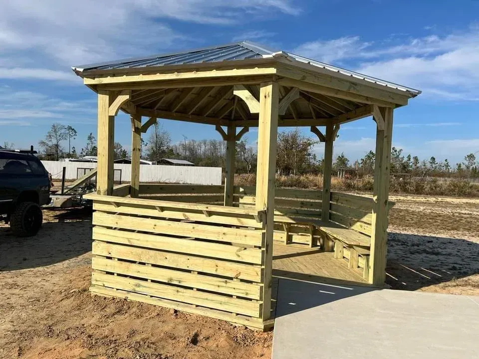 Wooden outdoor gazebo with slatted walls and a solar-panel roof on a dirt lot under a blue sky