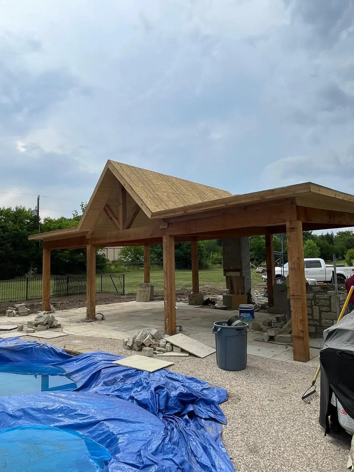 Wooden pavilion under construction with a peaked roof, blue tarp, and construction materials on site