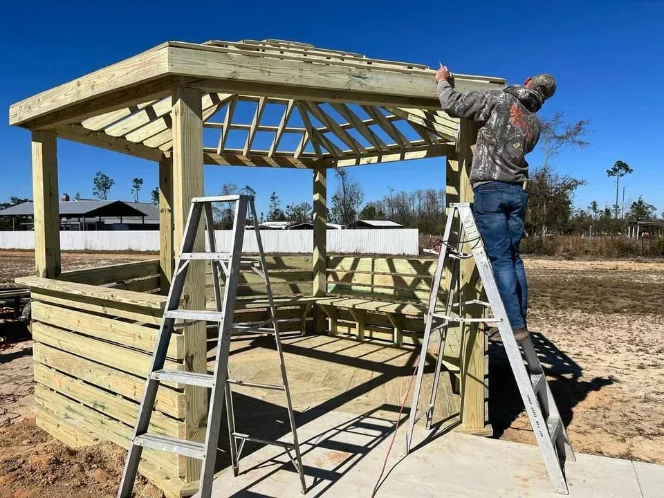 Worker installing roof on wooden gazebo frame beside ladder at a construction site