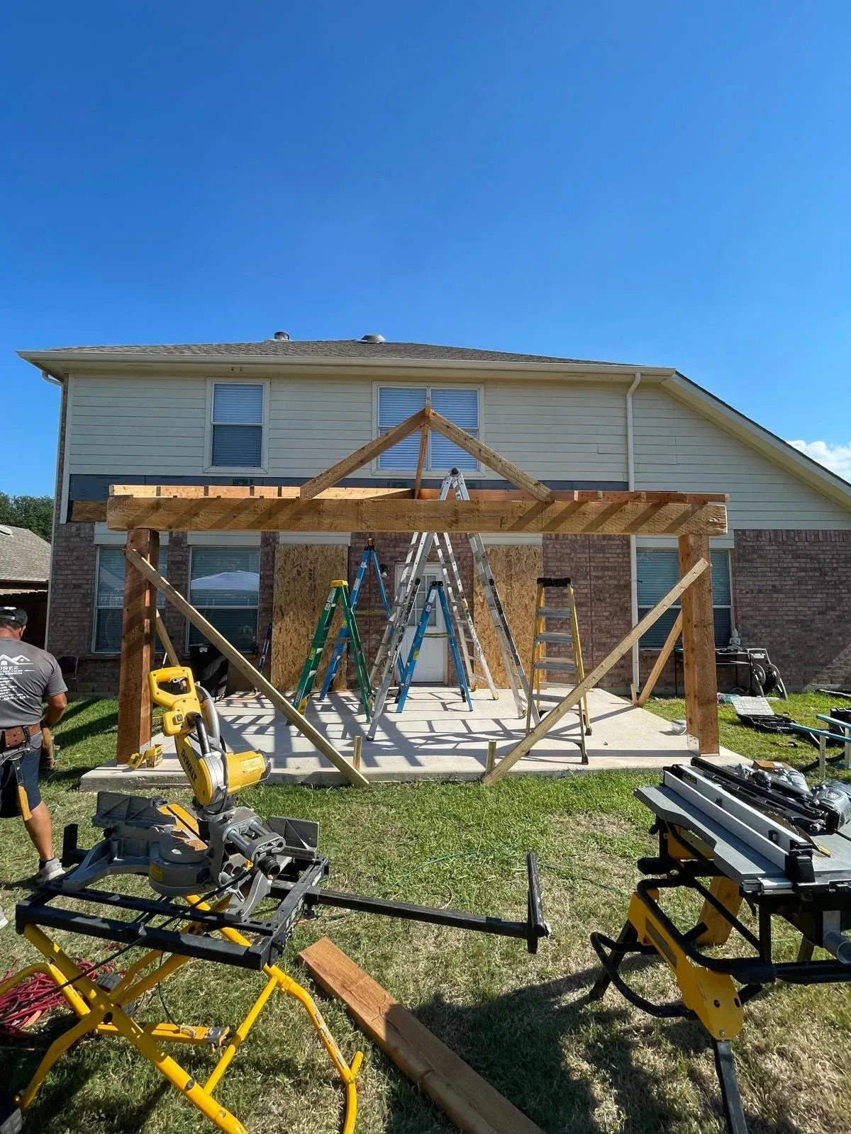 Workers build a wooden porch frame on the front of a two-story house under a clear blue sky.