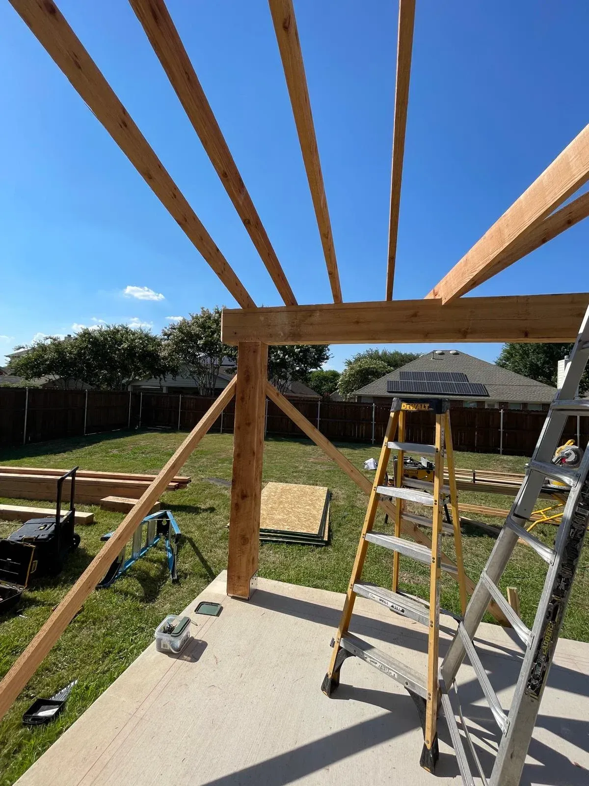 Backyard deck under construction with wooden beams, ladders, tools, and a blue sky