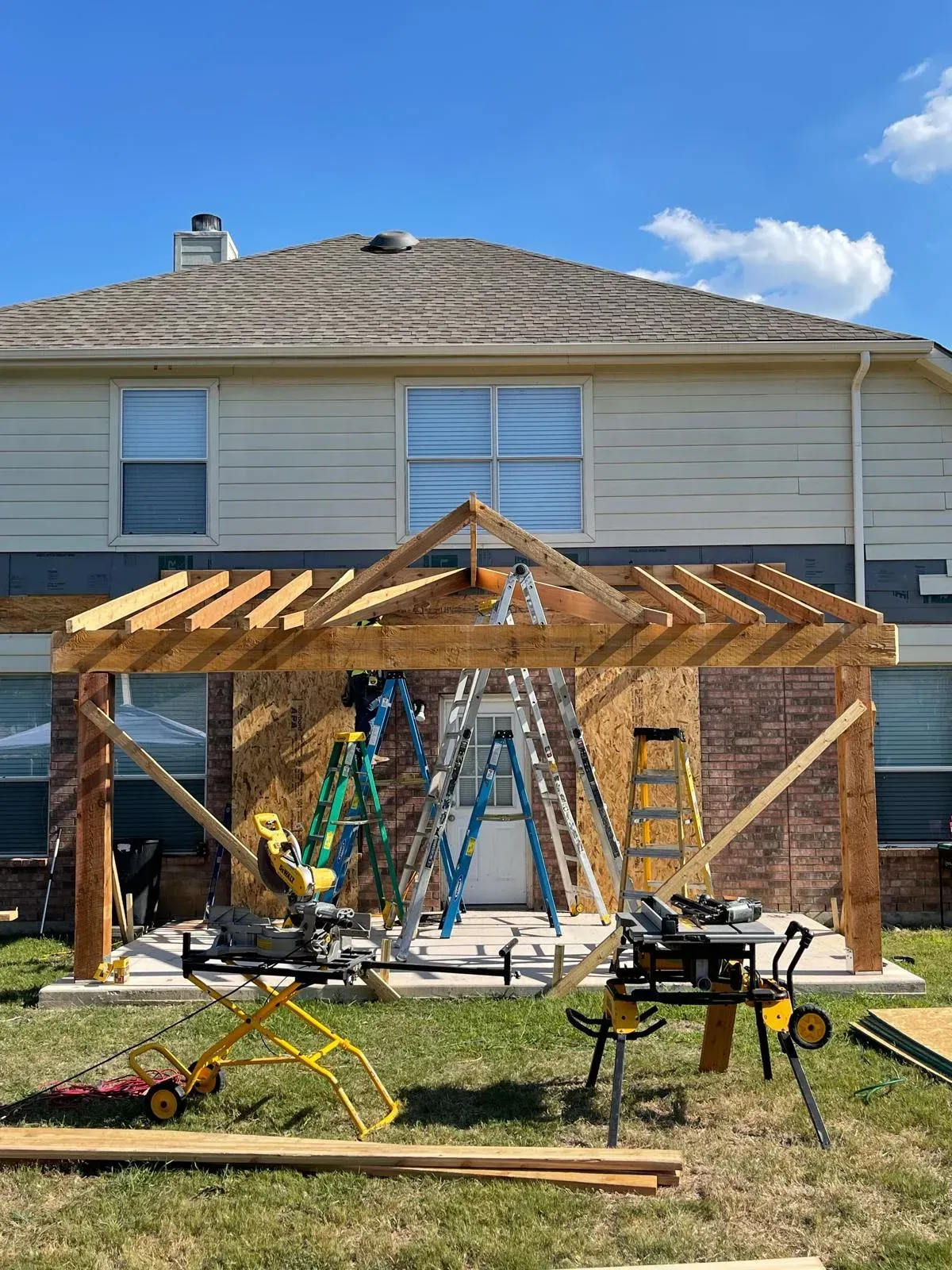Backyard pergola under construction beside a house, with tools, ladders, and exposed wooden framing.