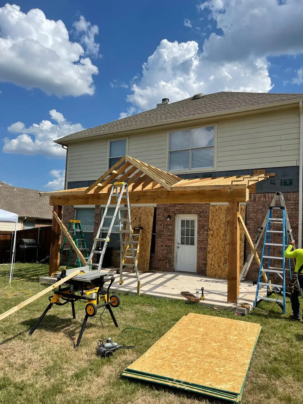 Backyard house under construction with ladders, roofing work, and a workbench under a partly cloudy sky