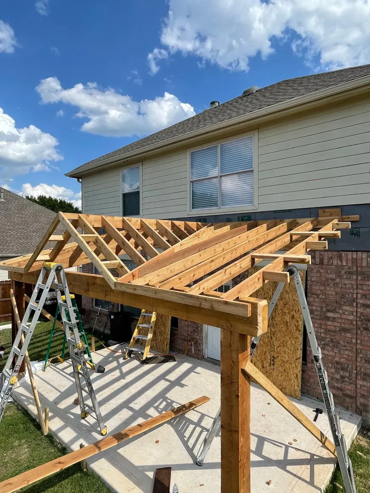 Partially built wooden patio roof frame beside a house under a blue sky