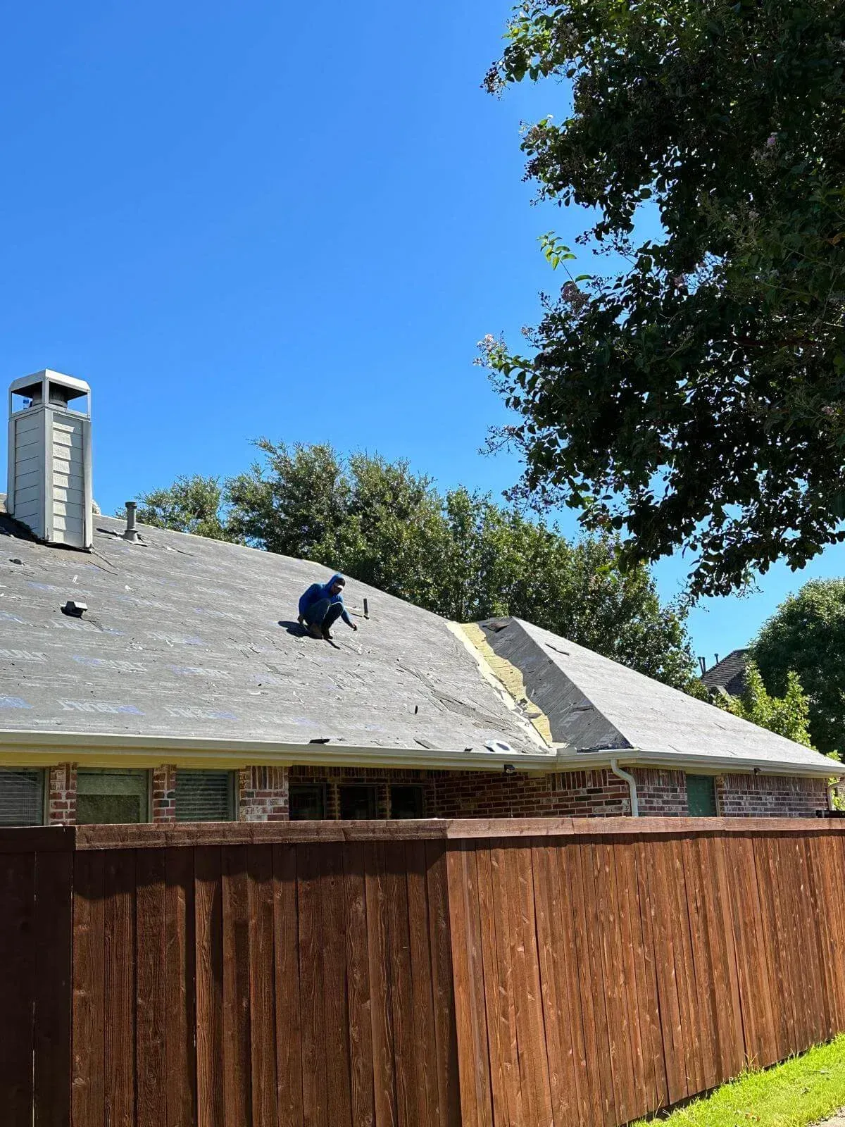 Worker on a sloped roof in a suburban backyard, with a wooden fence and clear blue sky.