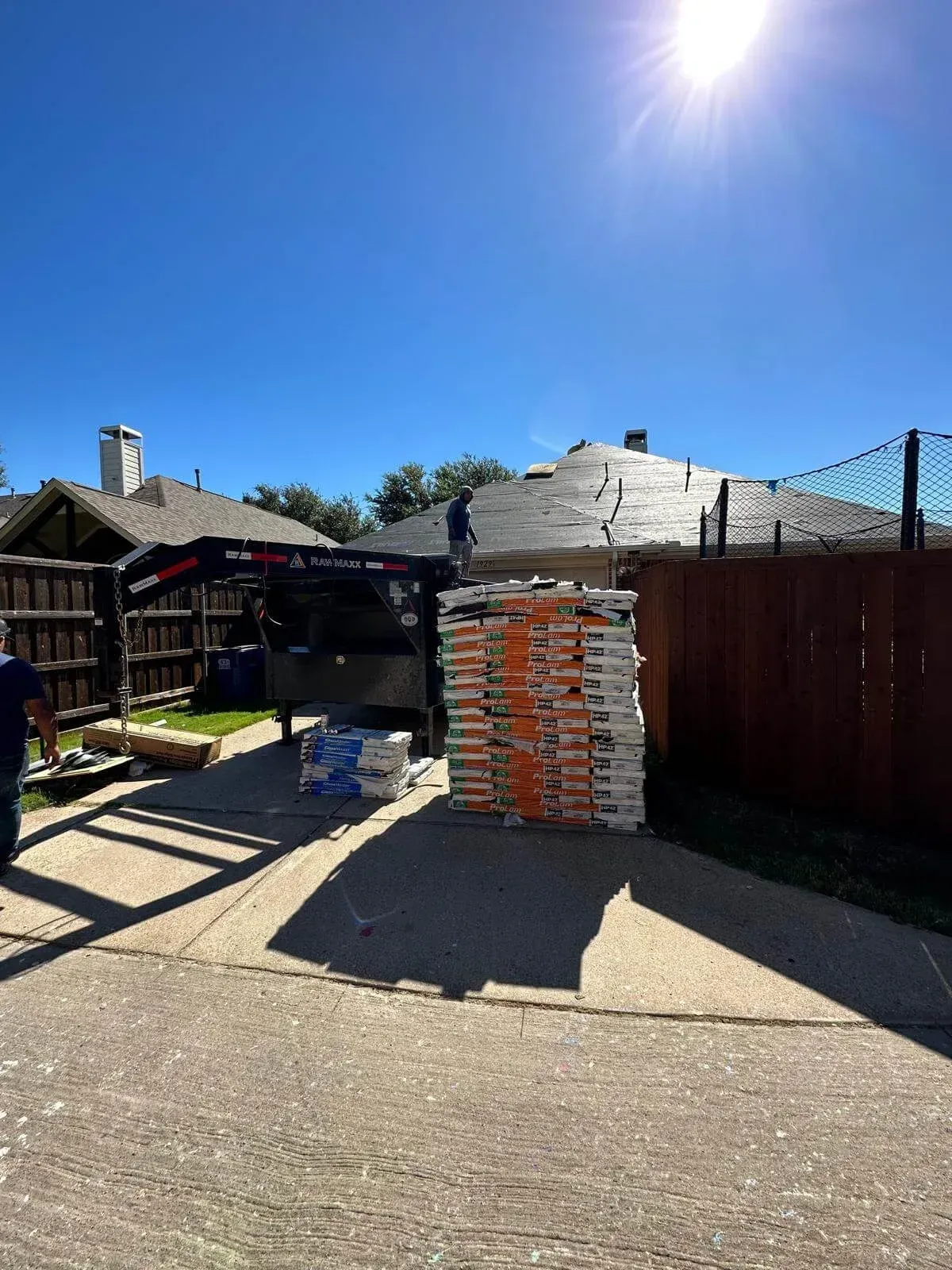 Backyard driveway with stacked bricks, a wooden fence, and bright sun overhead