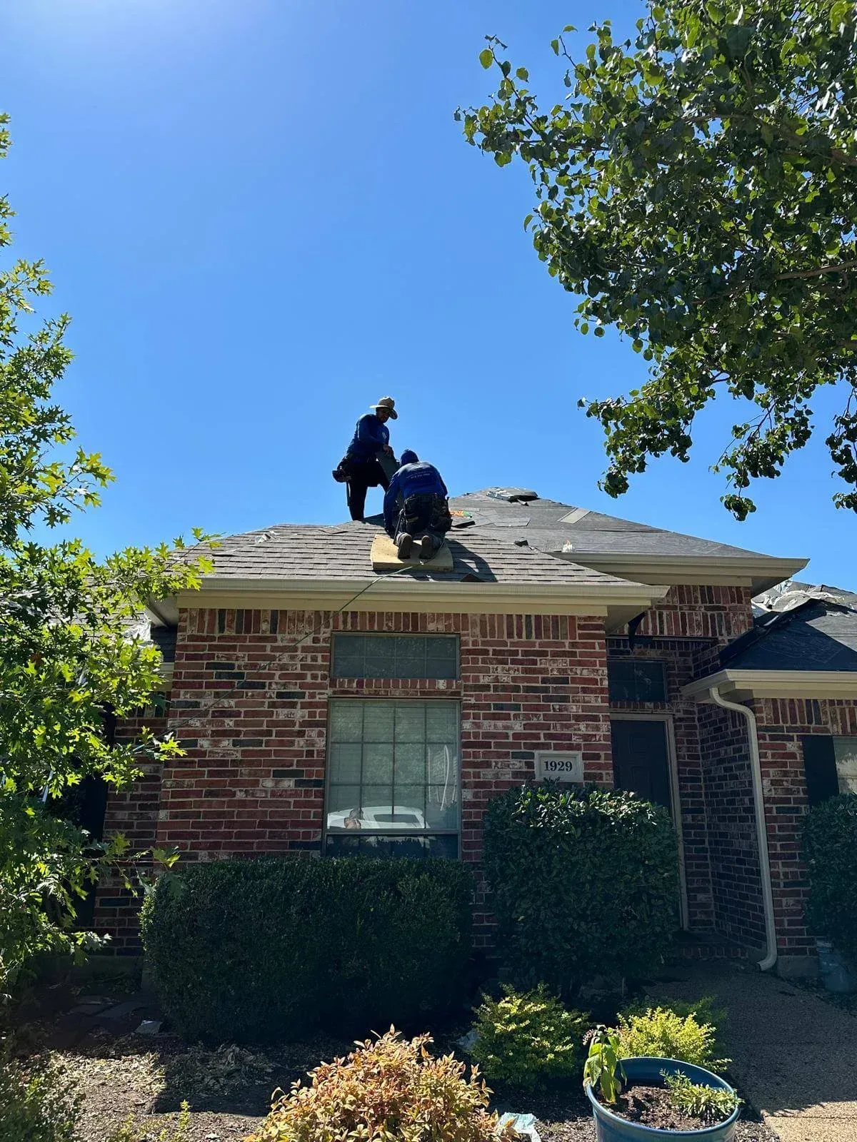 Two workers repairing a shingled roof on a brick house under a clear blue sky.