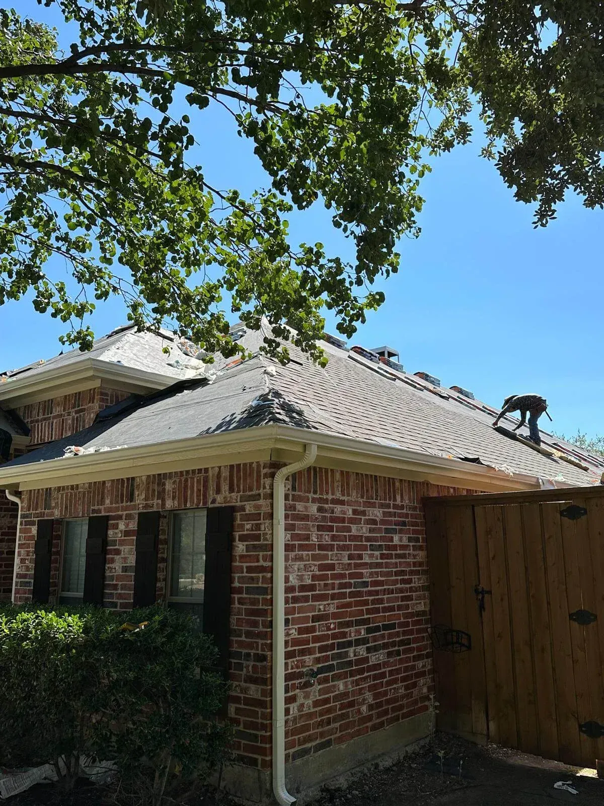 Brick house with a gray tiled roof, wooden gate, and leafy tree branches overhead on a sunny day