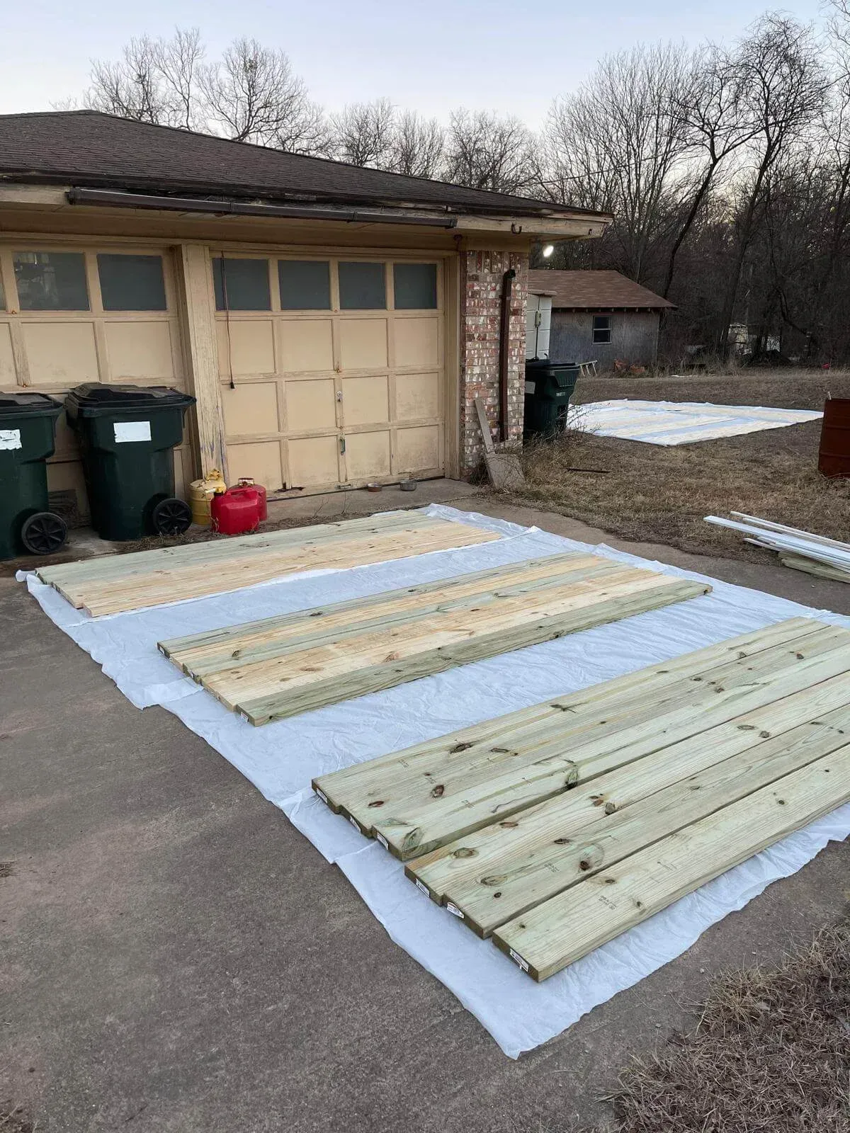 Wooden planks laid out on white tarp in a driveway beside a garage.