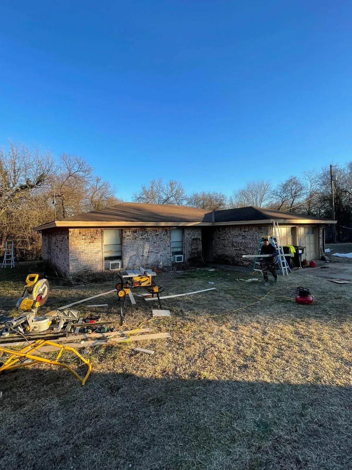 Single-story house with damaged front yard, debris scattered, and workers near the entrance under a clear blue sky