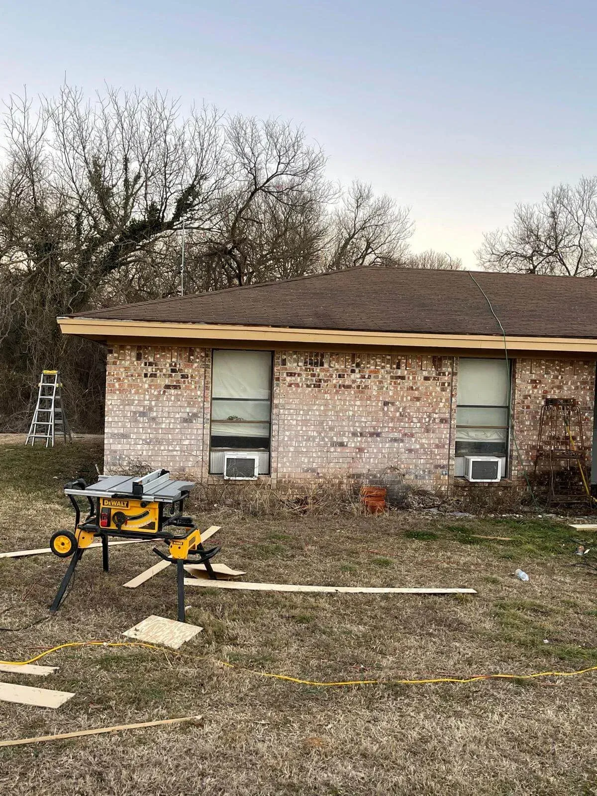 Single-story brick house with boarded window, tools, and ladder in a yard with scattered lumber