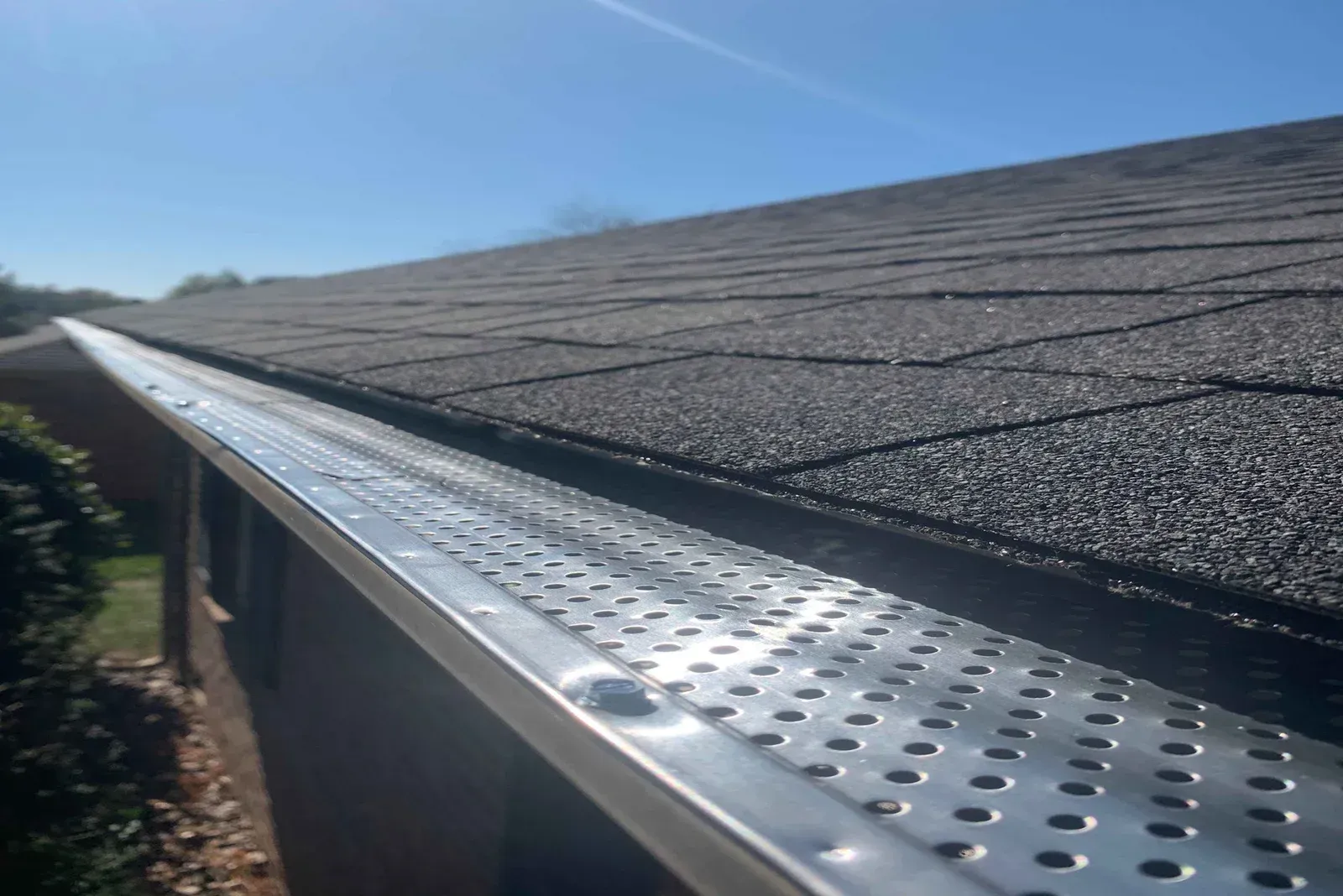 Close-up of a roof edge with a silver gutter guard under a shingled roof in bright sunlight