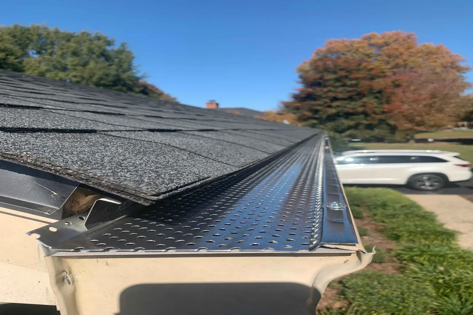 Close-up of a house roof edge with black shingles, white gutter, and a parked SUV in the background