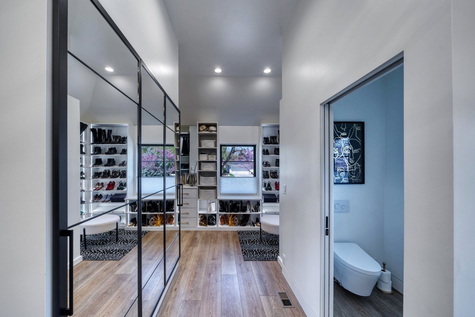 Modern walk-in closet with mirrored cabinets, shelves, wood floor, and an open doorway to a bathroom.