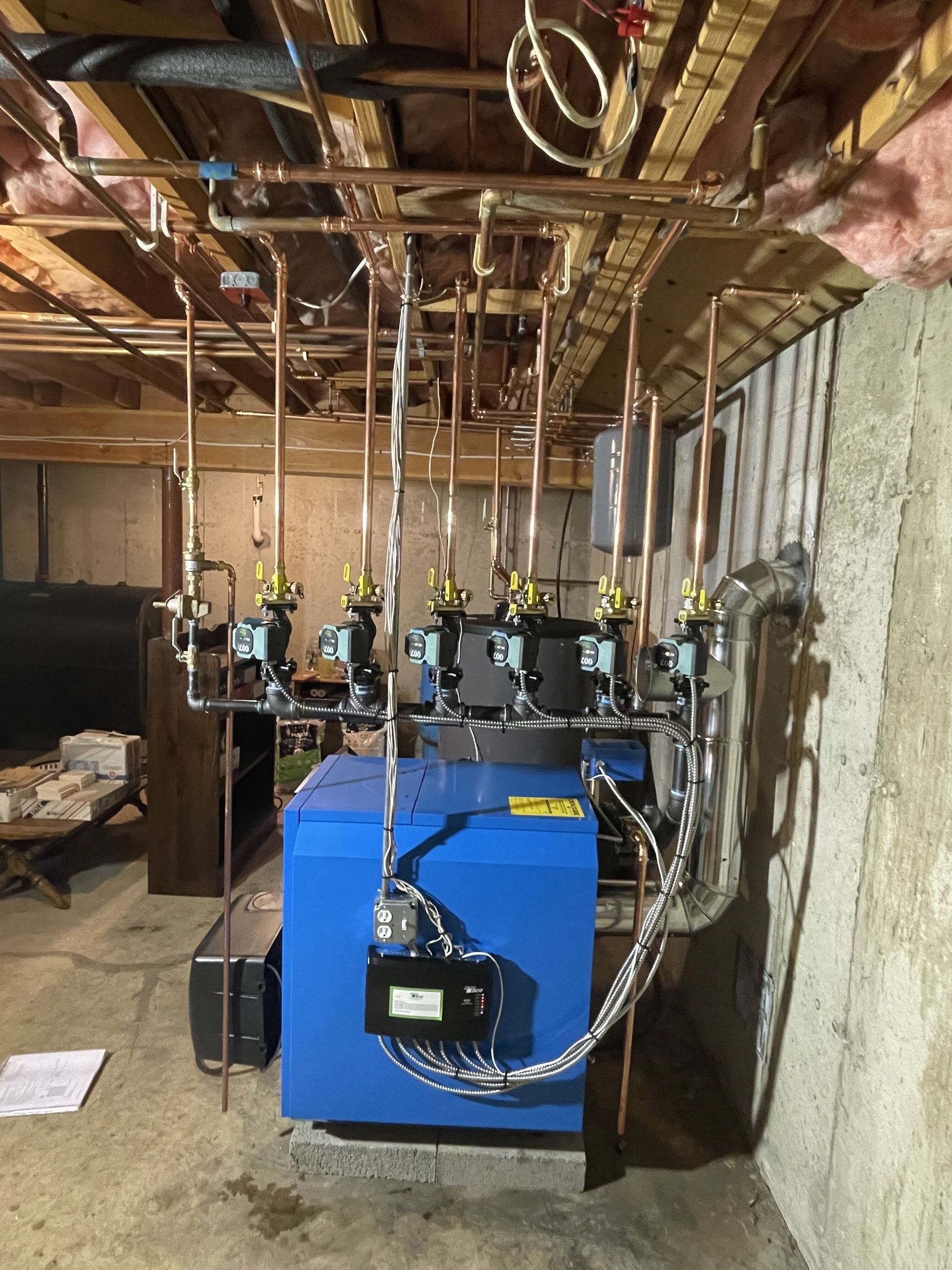 A blue boiler is sitting in a basement with pipes hanging from the ceiling.