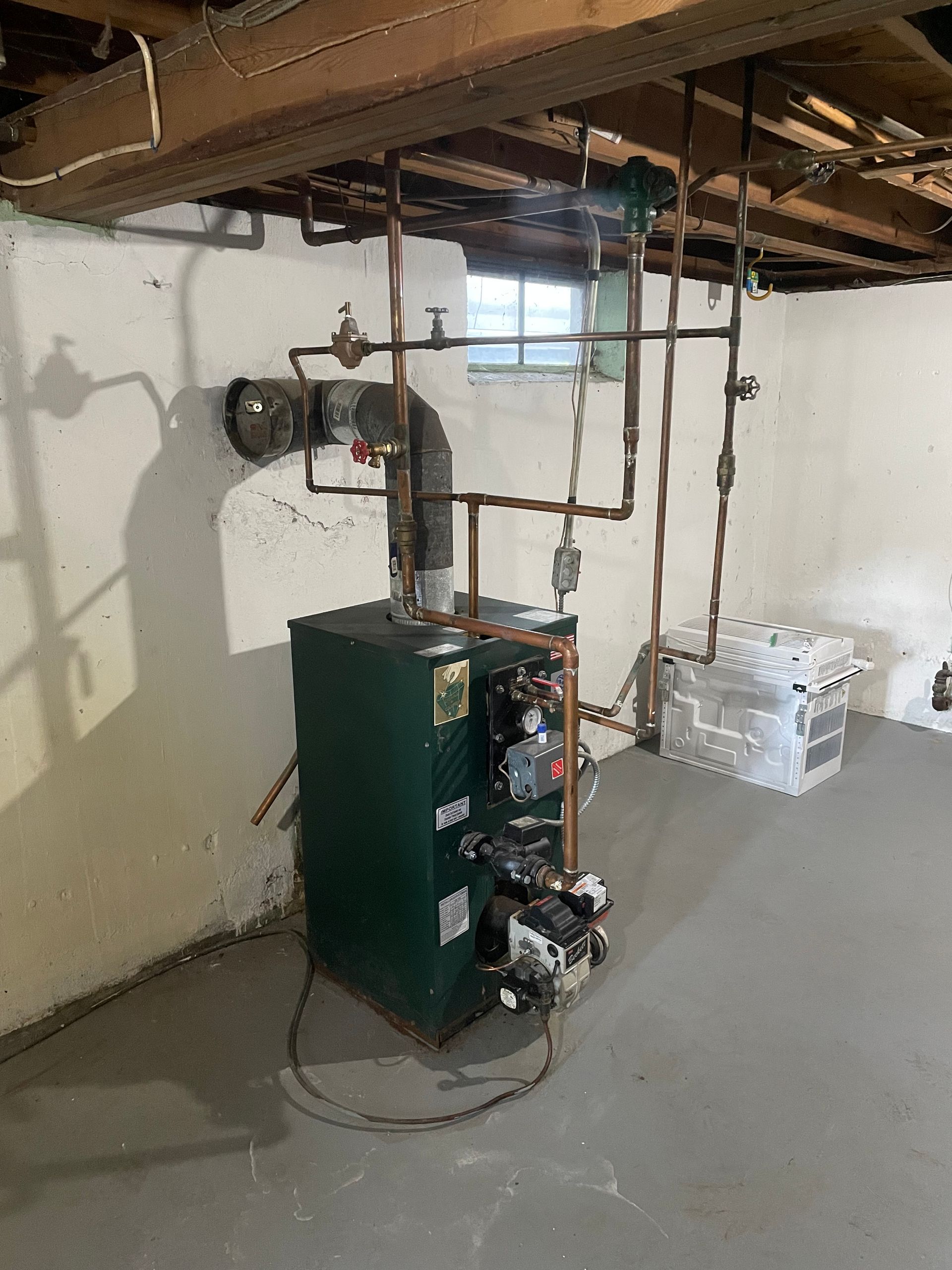 A green boiler is sitting in a basement next to a window.