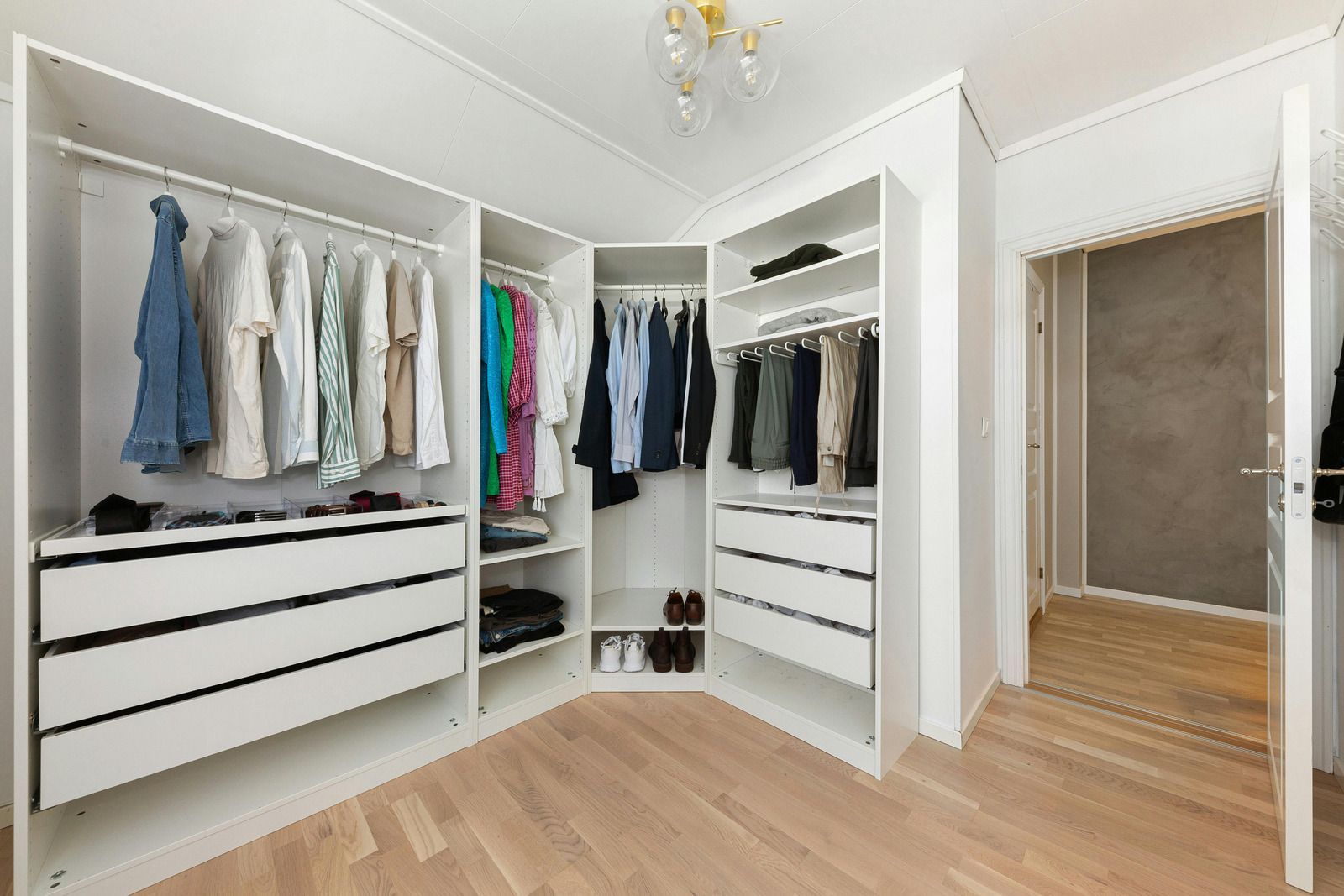 White walk-in closet with hanging clothes, drawers, and shelving. Includes a partially open doorway and light wood flooring.