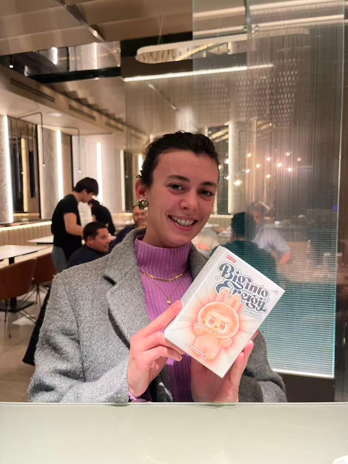 Una persona sorridente, seduta in un ristorante moderno, mostra una piccola scatola illustrata con l'immagine
