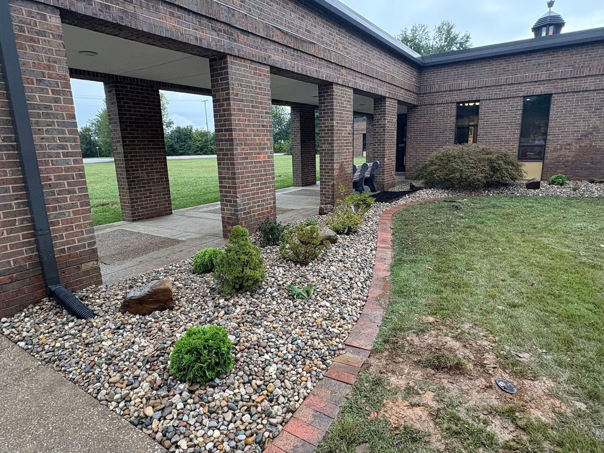 Brick courtyard with covered walkway, gravel bed, shrubs, and lawn beside a building