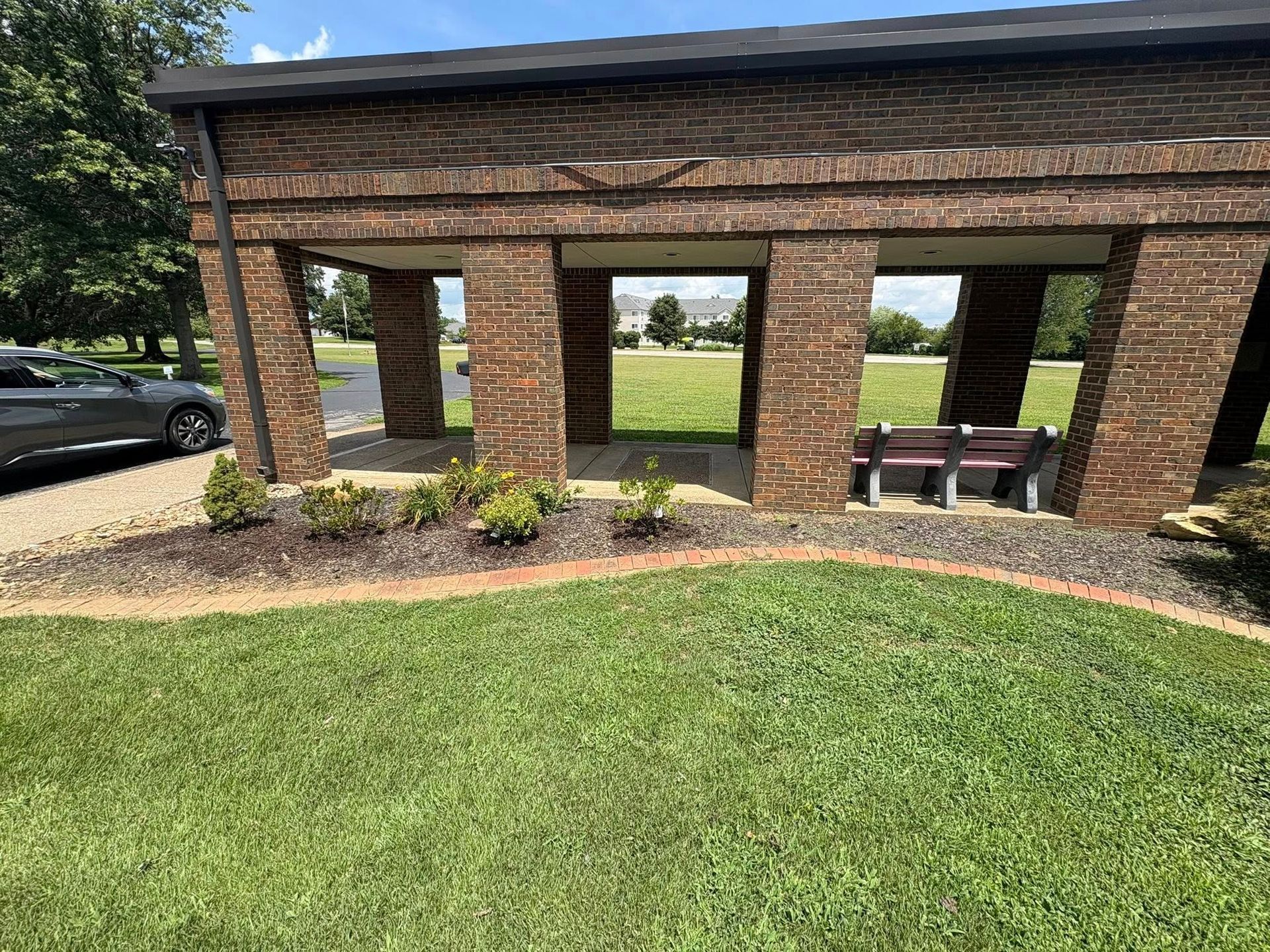 Brick pavilion with open windows, benches, and landscaped grass in a park area.