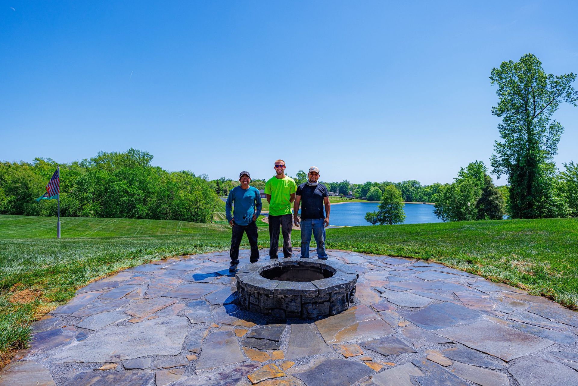 Three people stand by a stone fire pit on a lakeside patio under a blue sky.