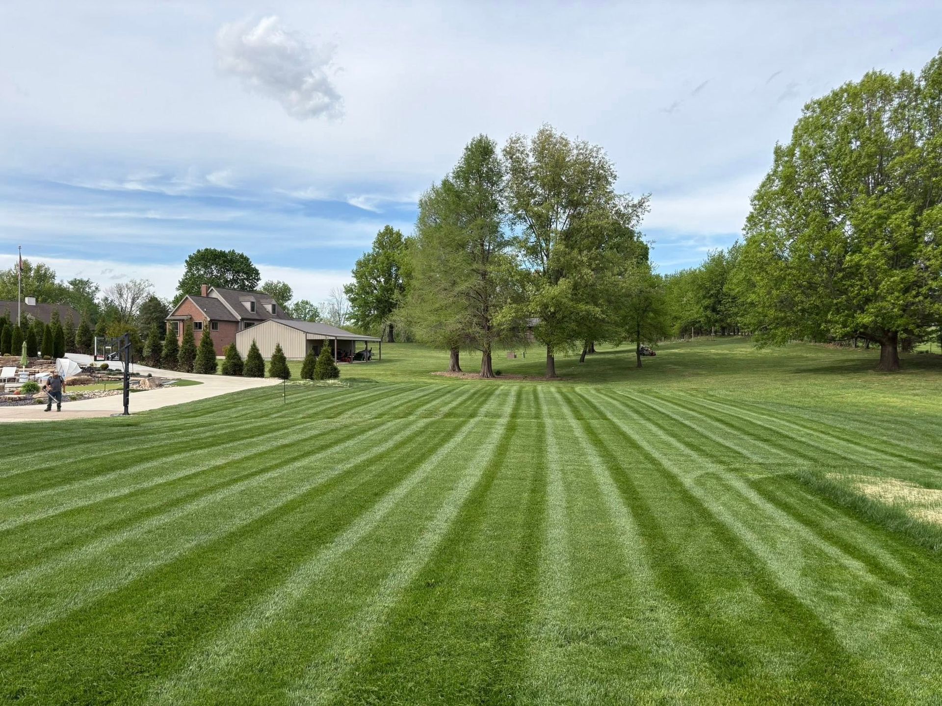 Wide green lawn with striped mowing lines, trees, and a small building under a cloudy sky.