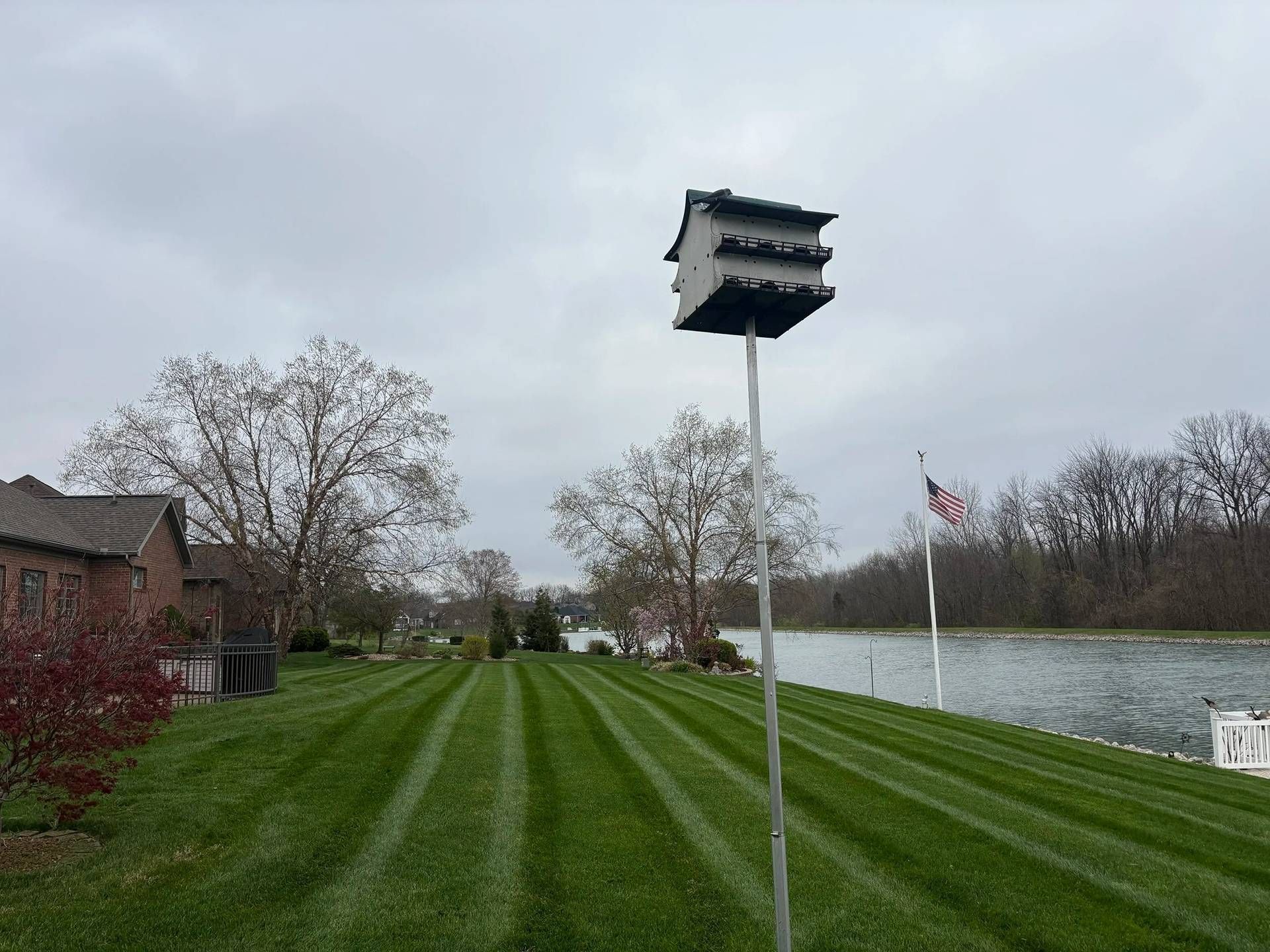 Grassy riverbank with a tall birdhouse, calm water, cloudy sky, and a house with a Canadian flag in the background
