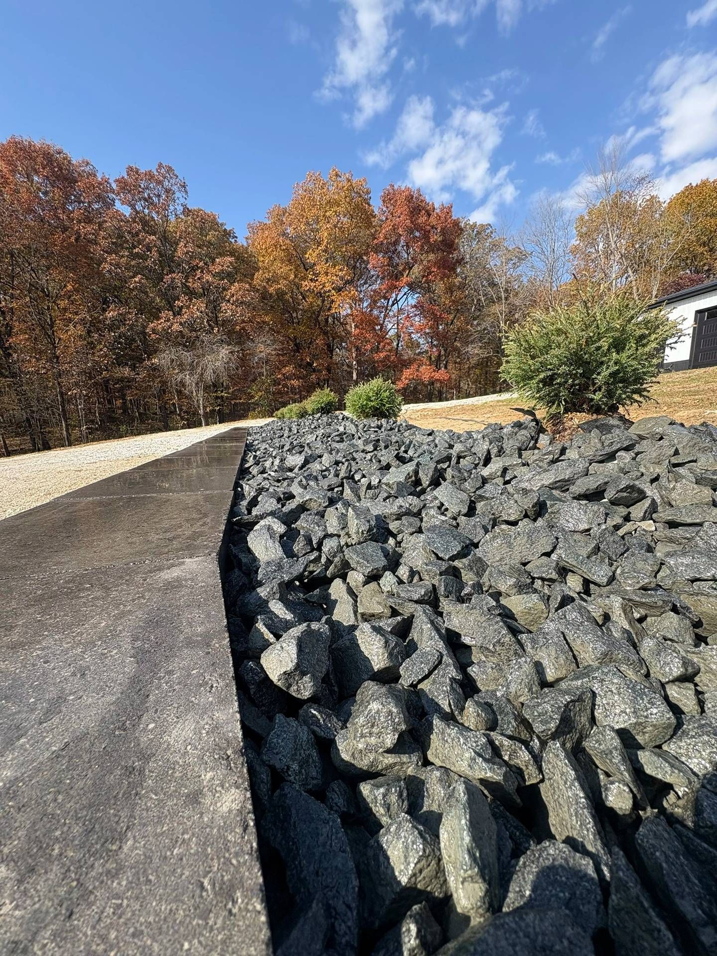 Paved path beside a gray rock retaining wall, with autumn trees and blue sky in the background