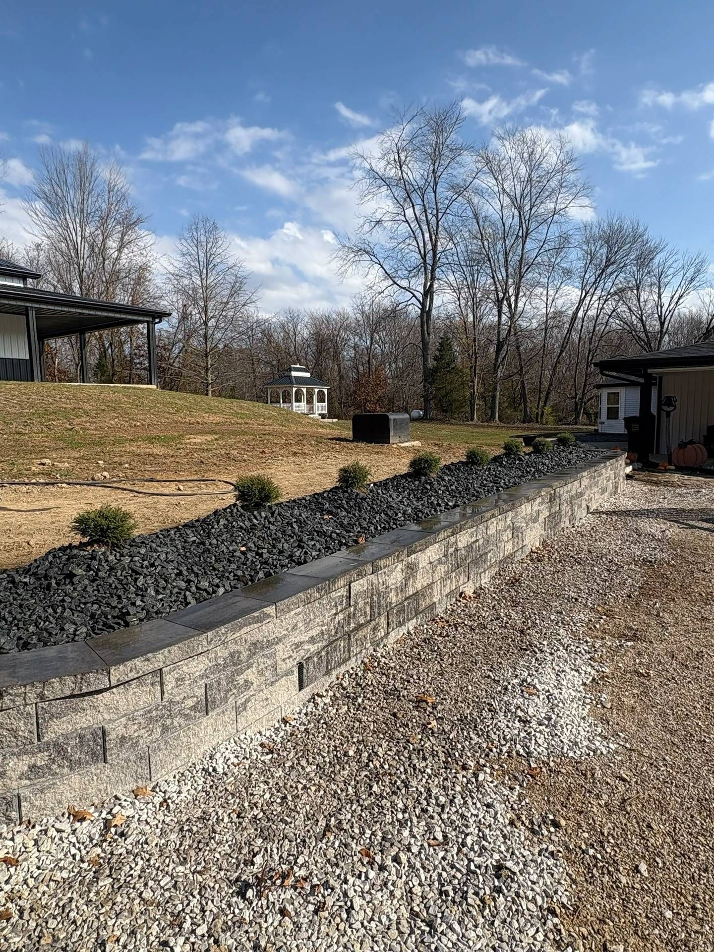 Gravel driveway beside a low stone wall and black mulch bed, with leafless trees under a blue sky.