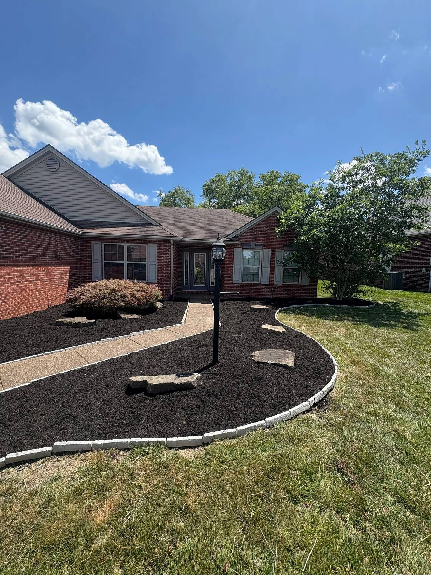 Brick house front yard with curved mulch bed, young tree, and blue sky