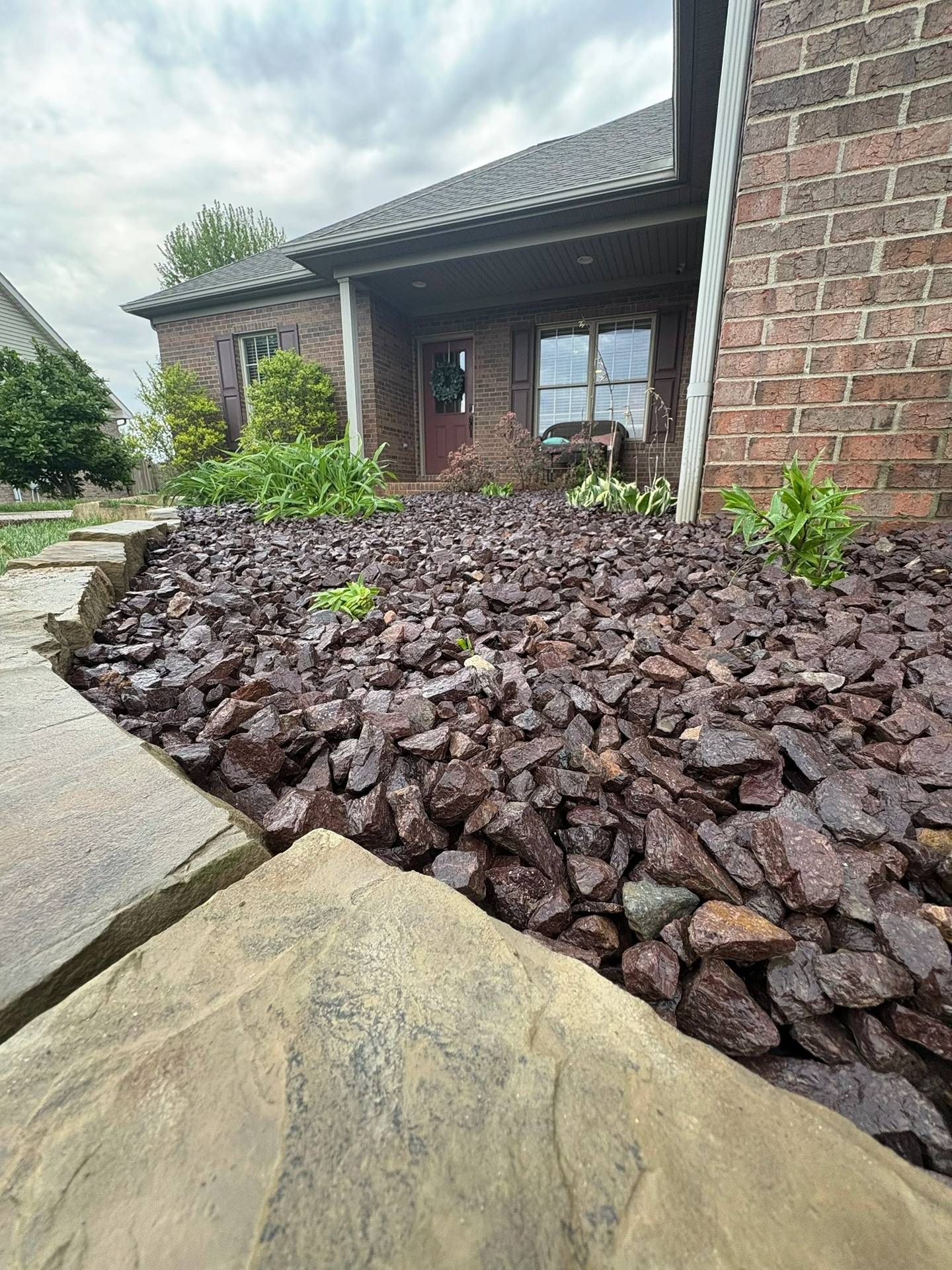 Front yard landscaping with dark stone mulch, small shrubs, and a brick house entrance under a cloudy sky