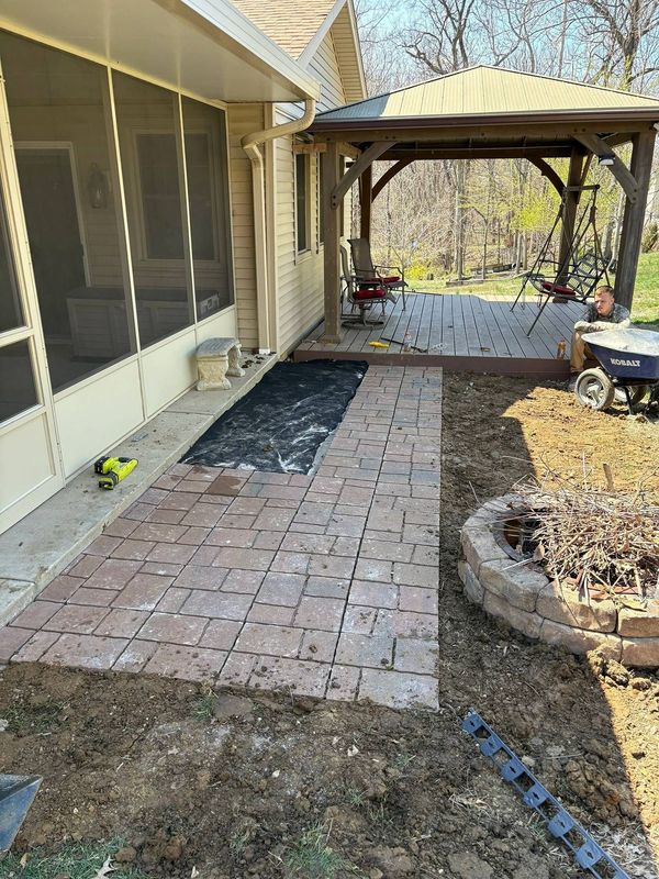 Brick patio walkway beside house leading to a wooden pergola with screened porch and garden beds.