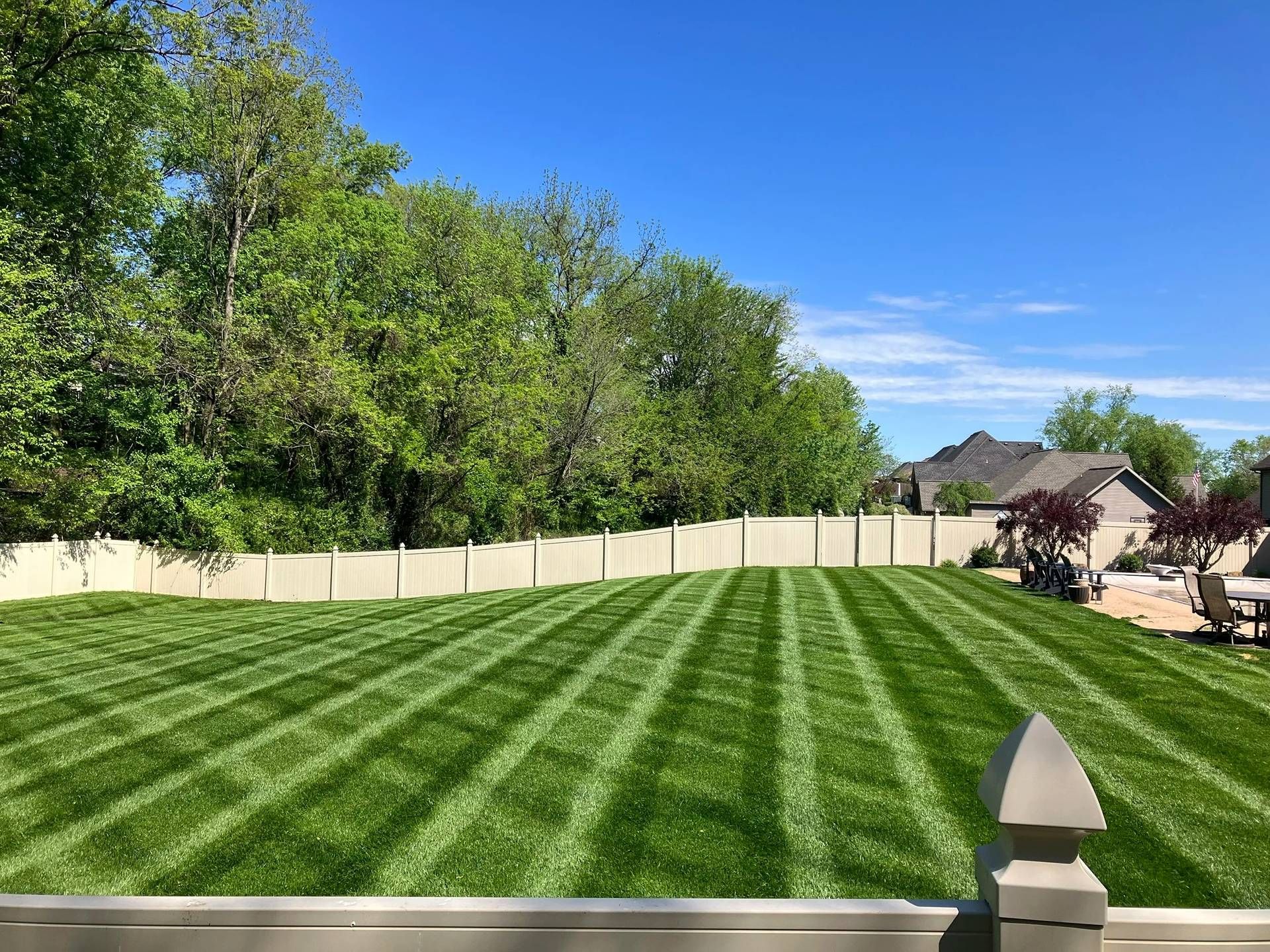 Freshly mowed striped lawn with a white fence, trees, blue sky, and a stone post in the foreground