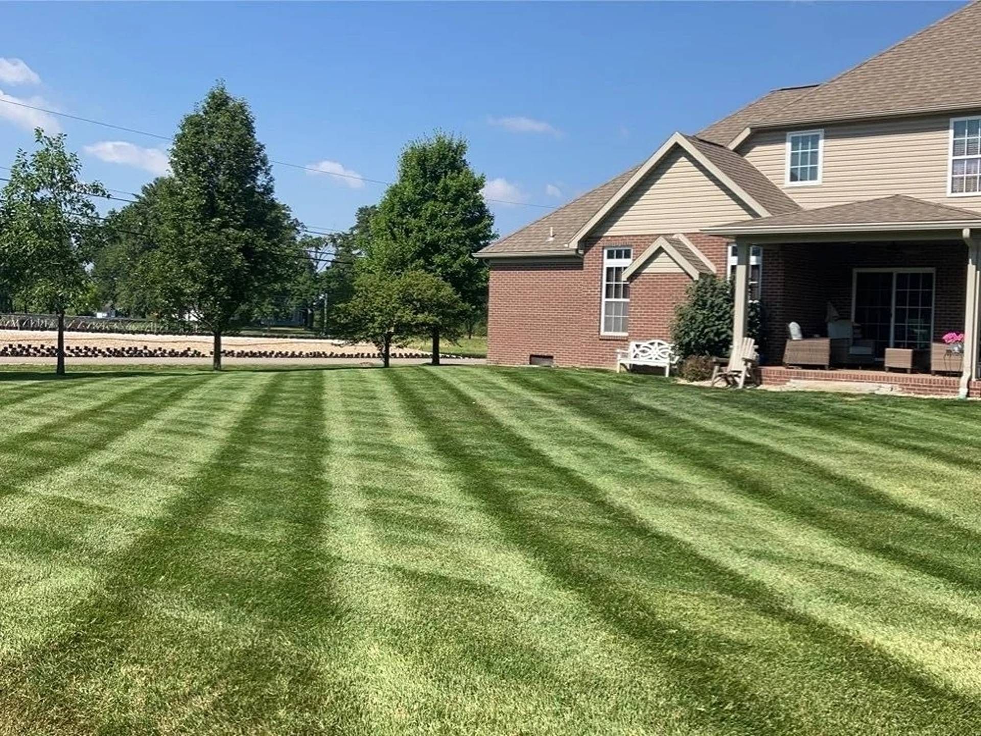 Backyard lawn with striped grass, trees, and a brick house with a patio on the right