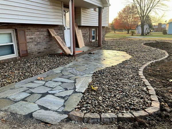 Stone walkway beside a house, curving through a gravel yard with brick edging and autumn trees.