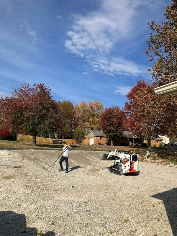 Person standing on a gravel lot with a small construction machine, autumn trees and blue sky in the background
