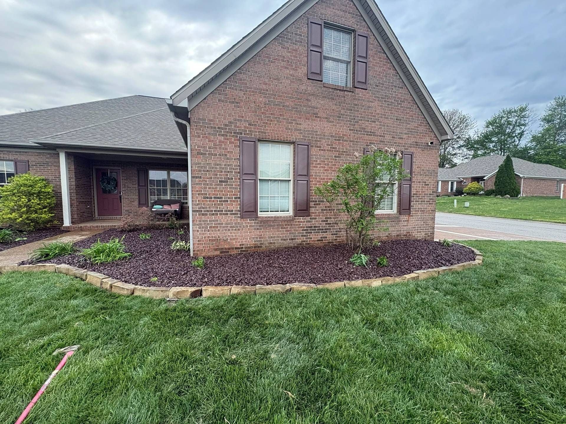 Brick house exterior with peaked roof, maroon shutters, and landscaped yard under cloudy sky