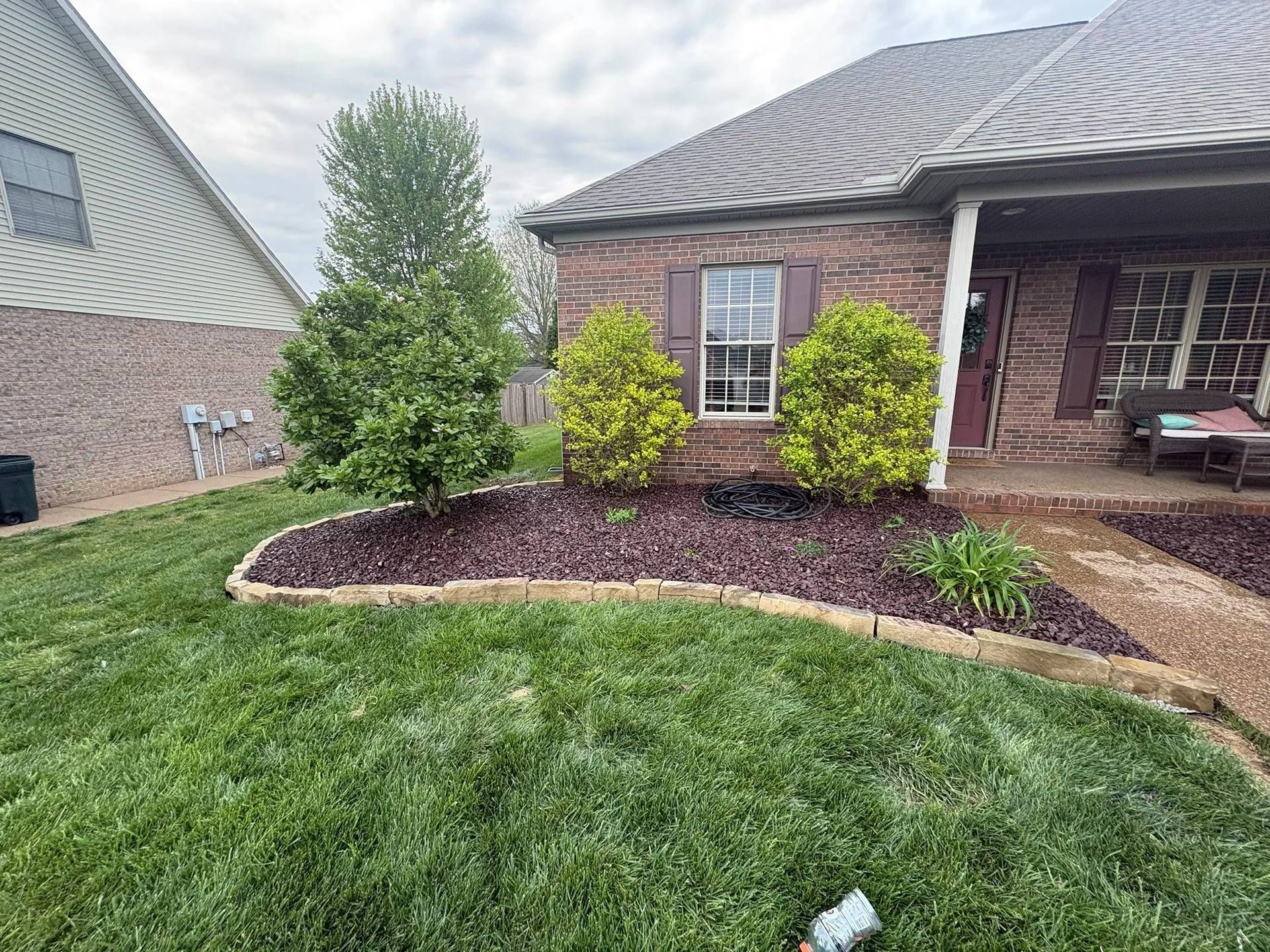 Front yard landscaping with green lawn, mulched garden bed, shrubs, and a brick house porch