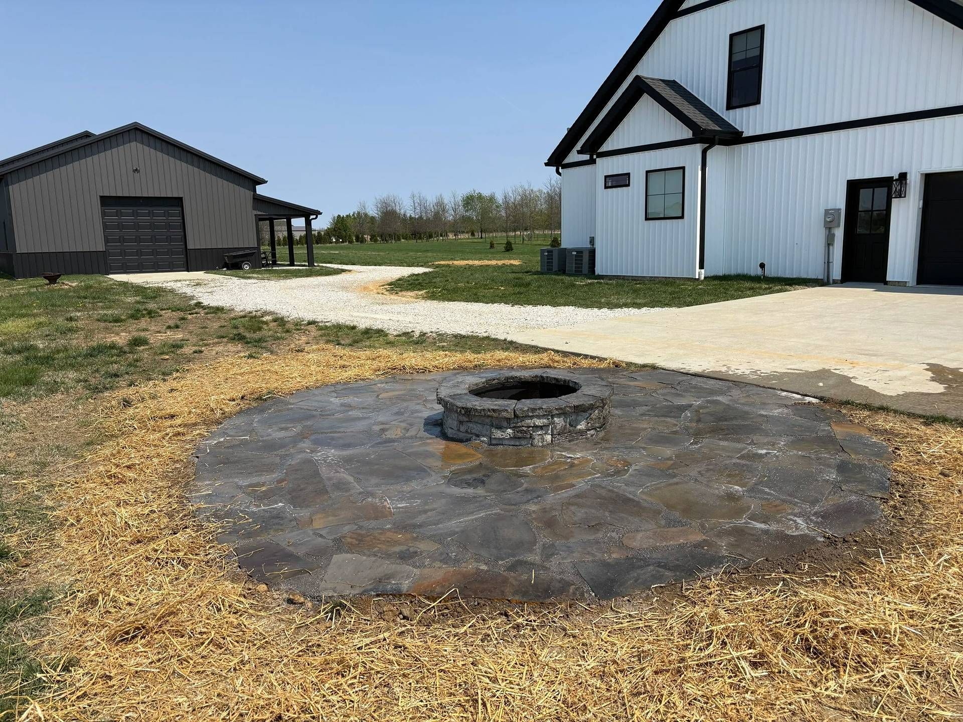 Rural yard with a circular fire pit in the foreground and two white-and-gray buildings in the background
