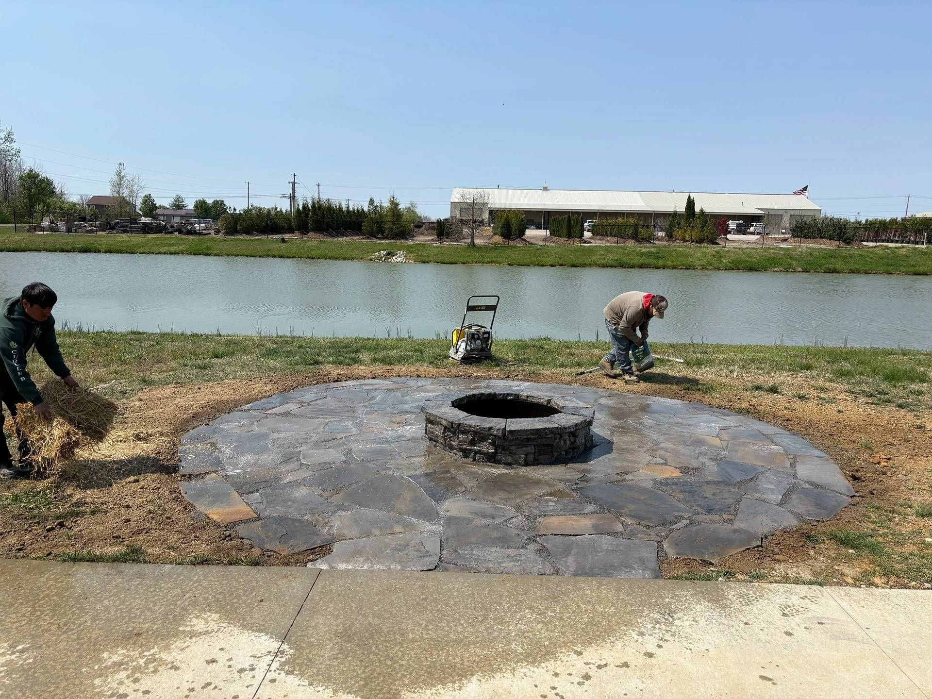 Workers landscaping around a circular stone fire pit beside a lake on a sunny day