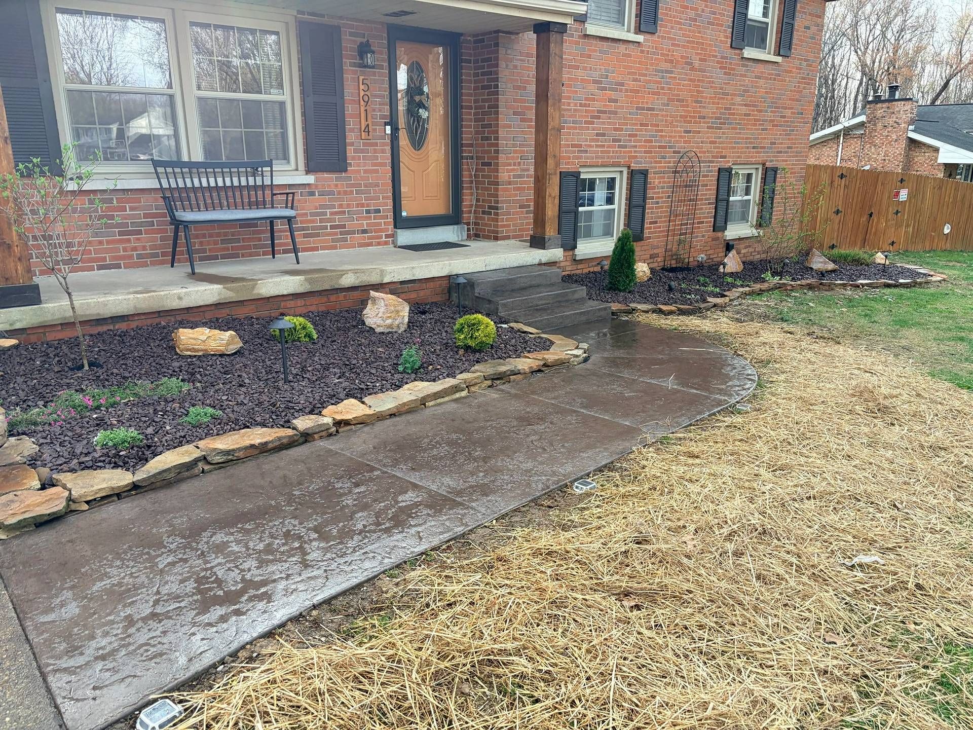 Front yard with brick house, wet concrete walkway, stone border, flower bed, and winter-yellow grass