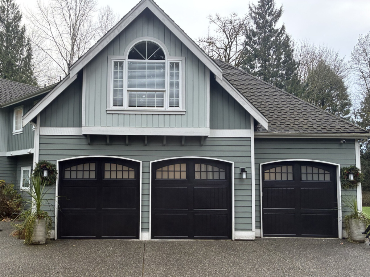 Gray house with three black garage doors and a gabled dormer, framed by trees