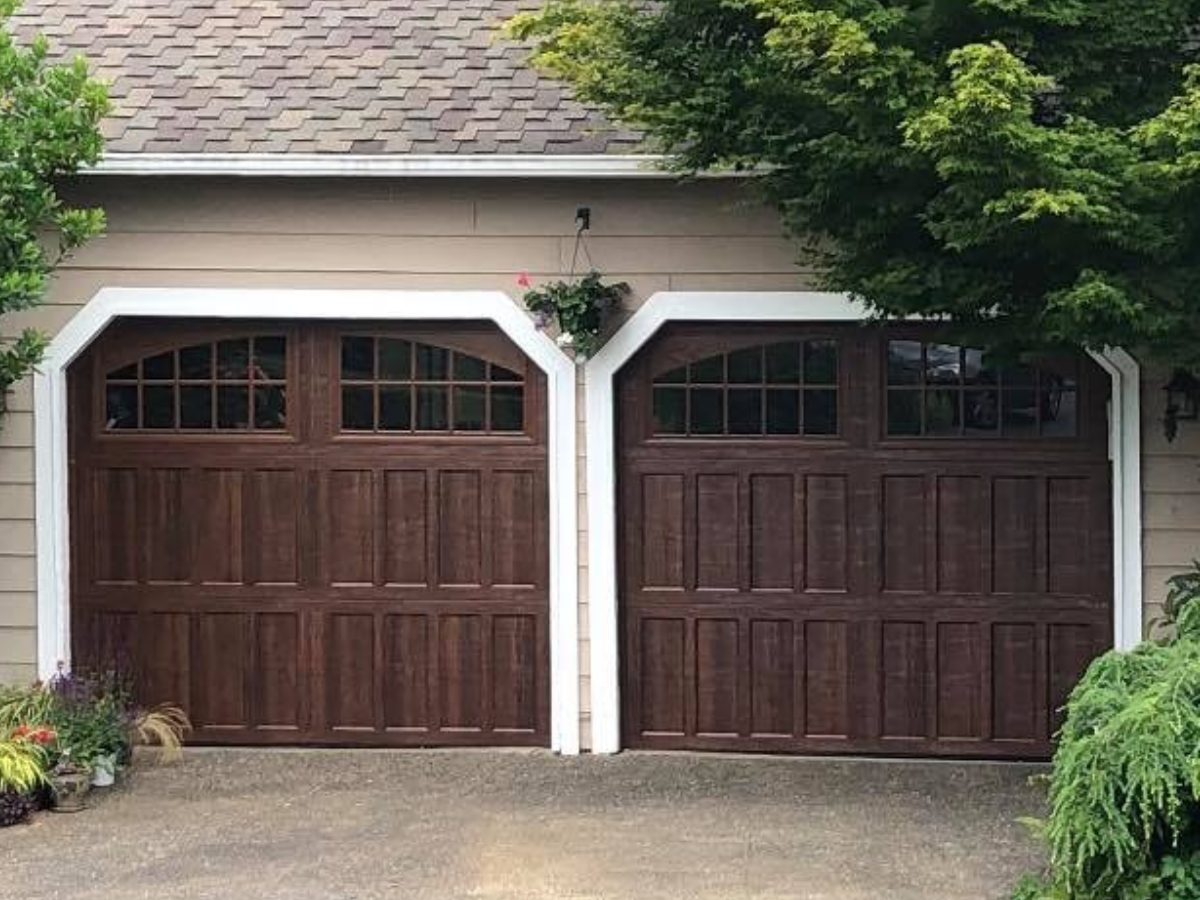 Two dark brown garage doors on a house with white trim and a shingled roof.