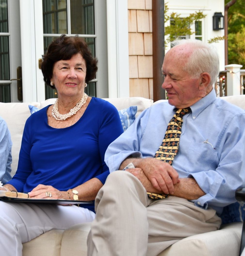 A couple sits together on an outdoor sofa; the person on the left wears a blue top, and the person on the right a tie.