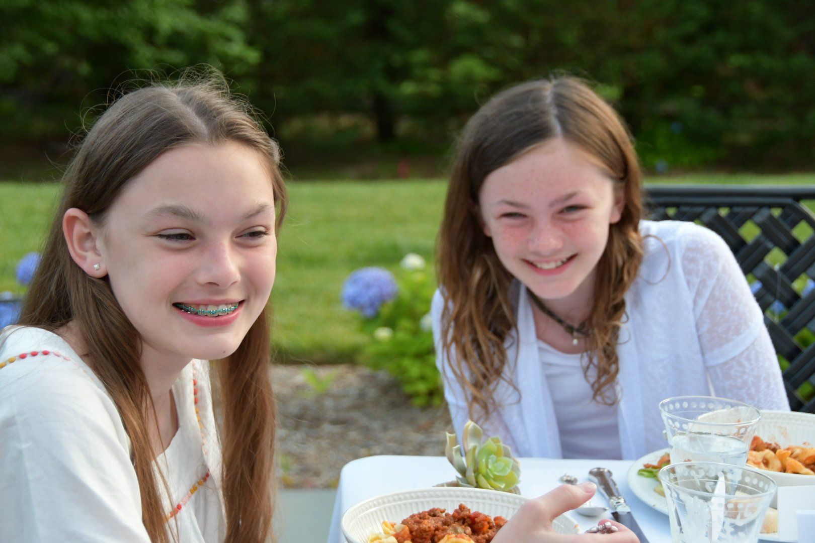 Two smiling individuals seated at an outdoor table with white plates of food, wearing white tops with greenery behind them.