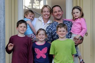A smiling family of eight stands in front of a light-colored doorway.