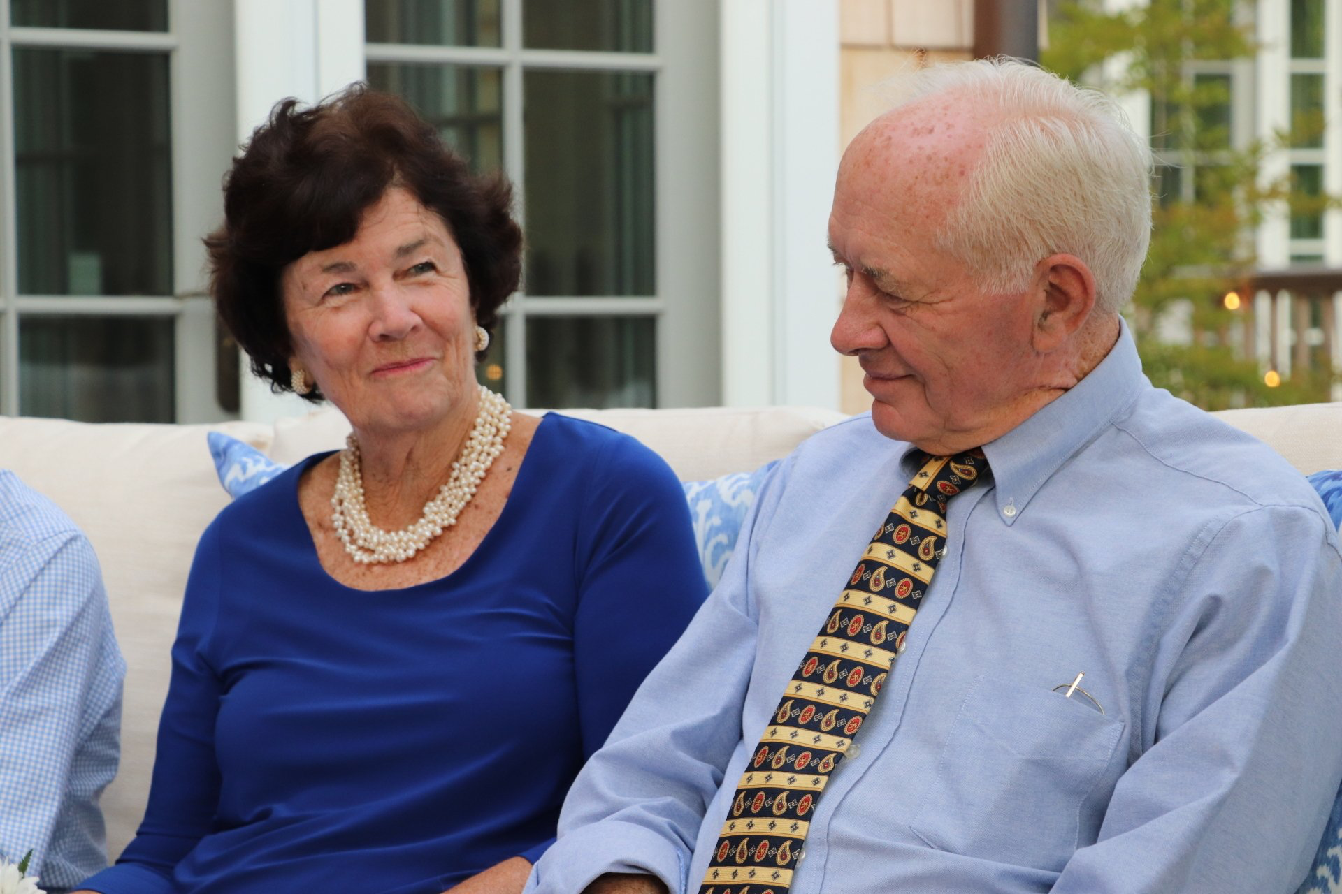 An older couple in blue attire sits outdoors, smiling at each other. The woman wears a pearl necklace.