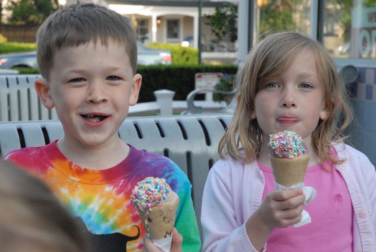Two children smile while holding ice cream cones topped with colorful sprinkles at an outdoor seating area.