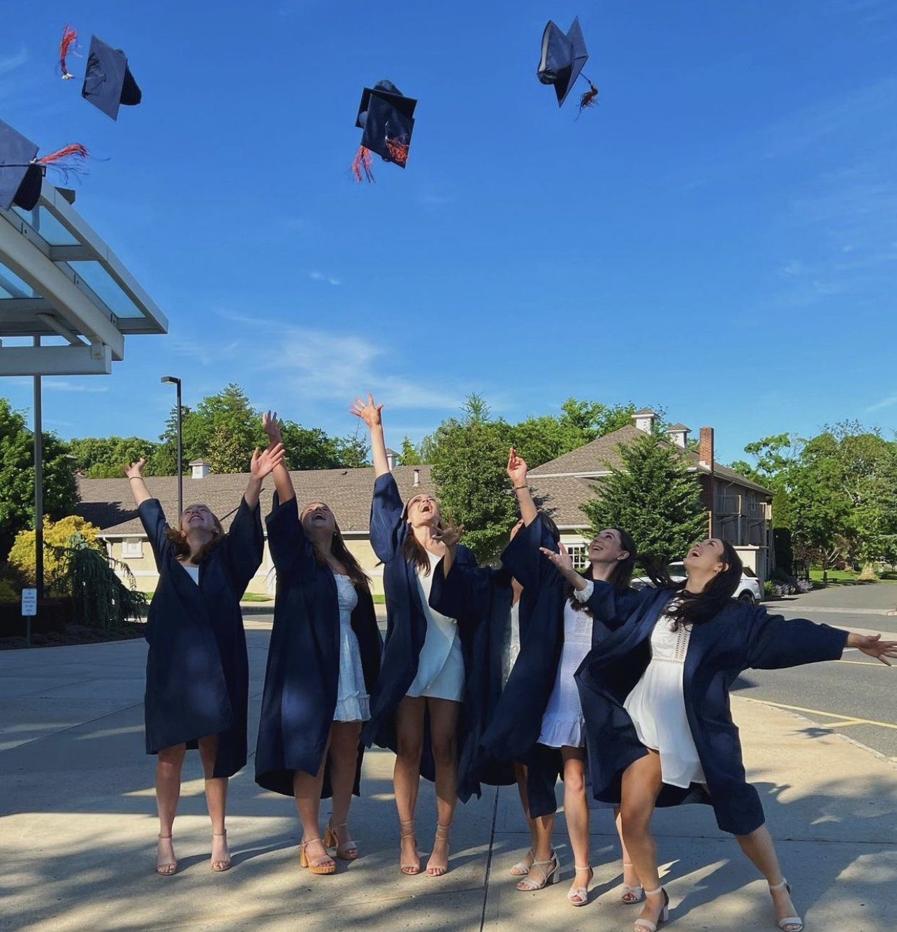 Six graduates in dark robes and white dresses toss their caps into the air outdoors on a sunny day.